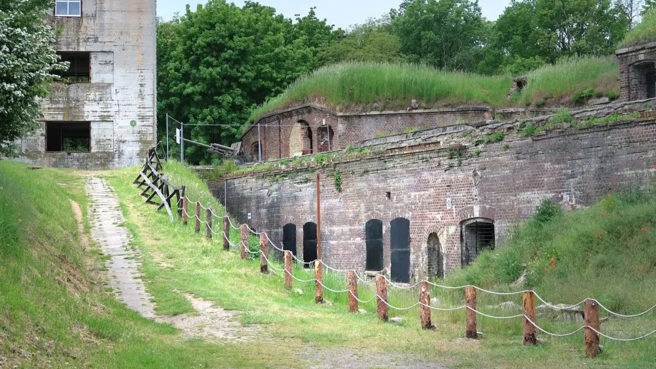 Fort Zachodni w Świnoujściu, zarośnięty trawą i otoczony zielenią, z widocznym starym budynkiem i historyczną artylerią.