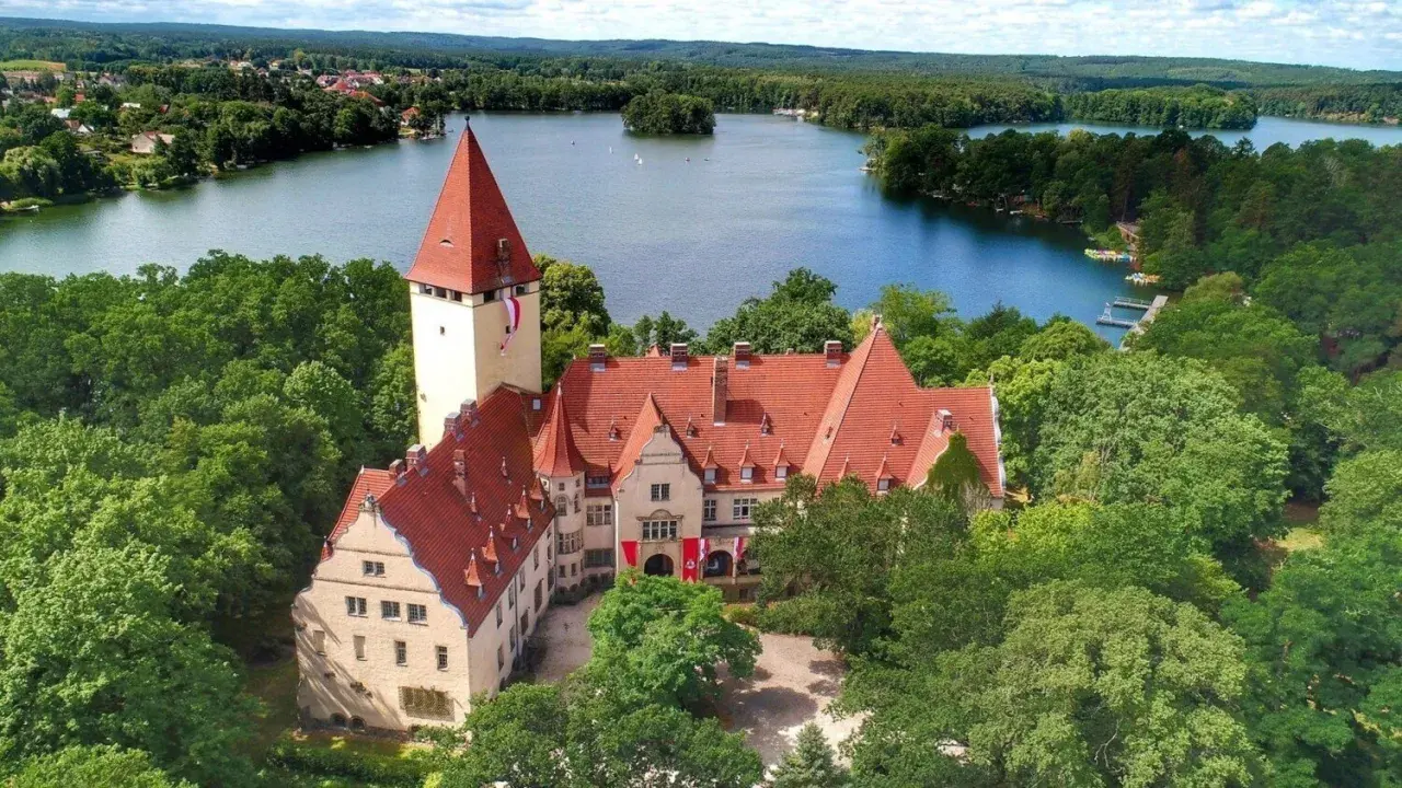 Zamek w Lubniewicach otoczony drzewami, nad jeziorem.