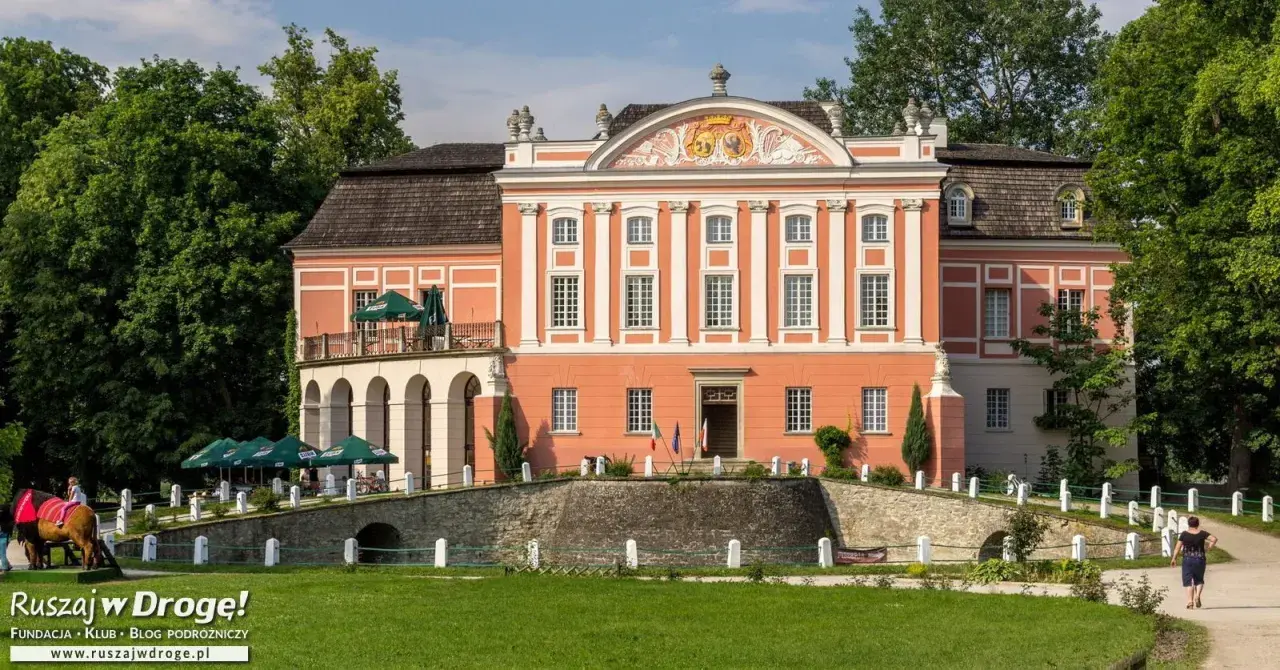 Kurozwęki pałac, z pomarańczową fasadą i białymi zdobieniami, otoczony zielenią. Na pierwszym planie mostek i trawnik.
