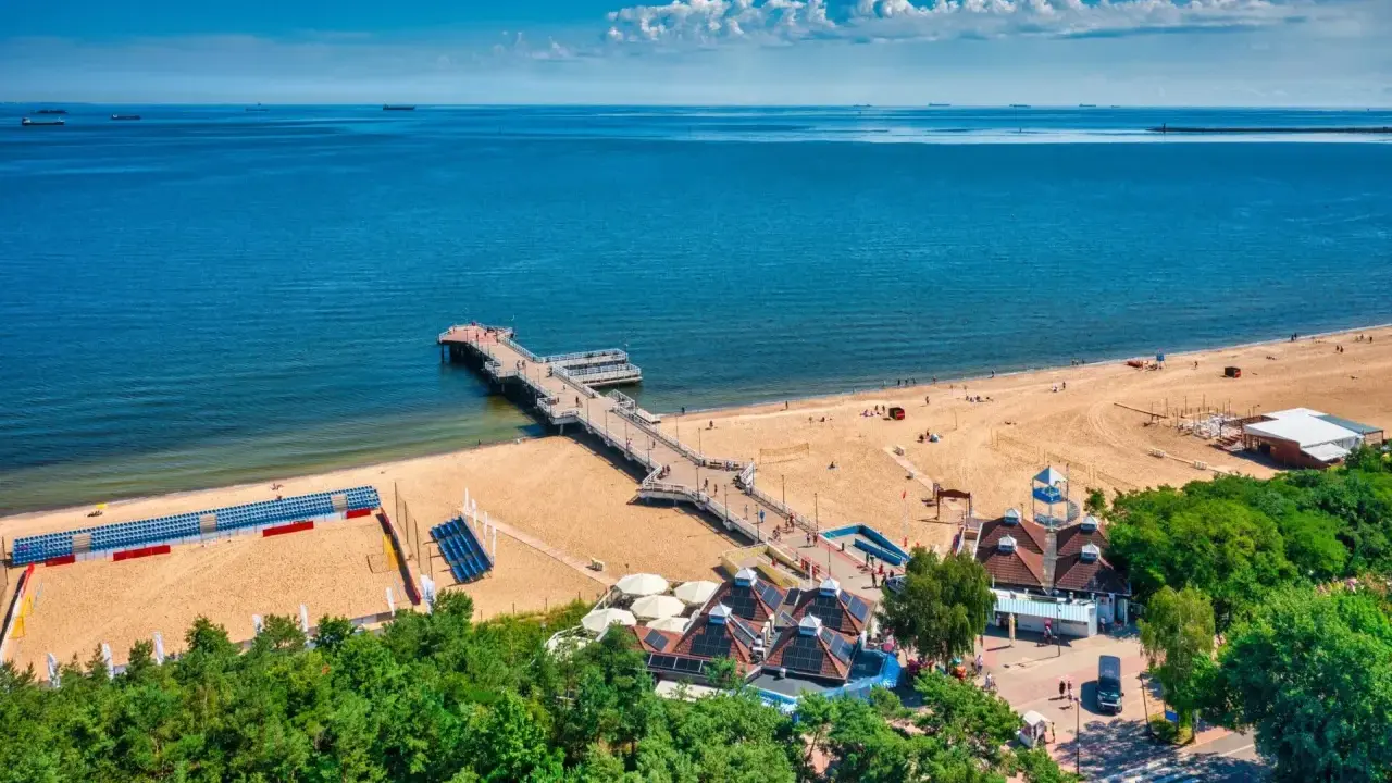 Idealne miejsce, gdzie na wakacje w sierpniu: piaszczysta plaża, molo, morze i zielone drzewa.