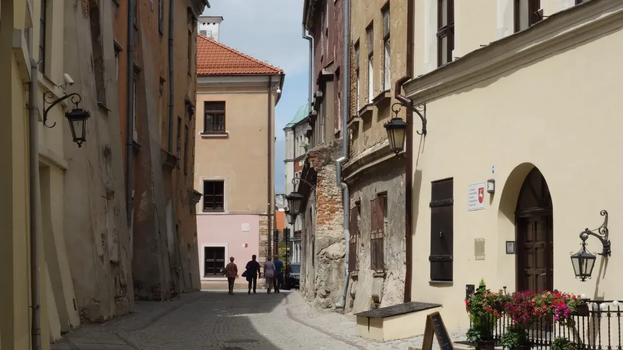 Wąska, brukowana uliczka w Lublinie z kamienicami i kilkoma spacerującymi osobami.