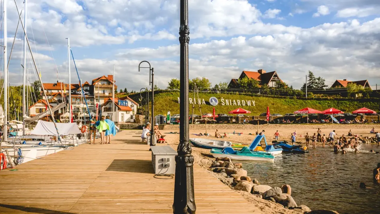 Słoneczna plaża nad jeziorem Śniardwy tętni życiem. Ludzie odpoczywają na piasku, kąpią się w wodzie, a obok cumują żaglówki.