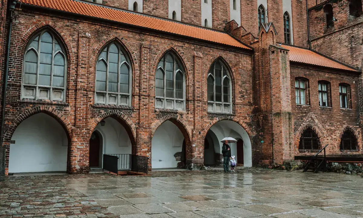 Zamek w Kwidzynie, z ceglaną fasadą i arkadowymi przejściami, w deszczowy dzień. Dwie postacie z parasolem spacerują po mokrym dziedzińcu.
