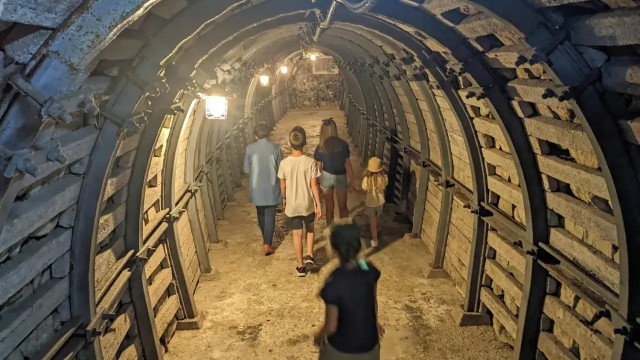 Rodzina zwiedza podziemia Sandomierz, idąc przez oświetlony tunel z kamiennymi ścianami i metalowymi wzmocnieniami.