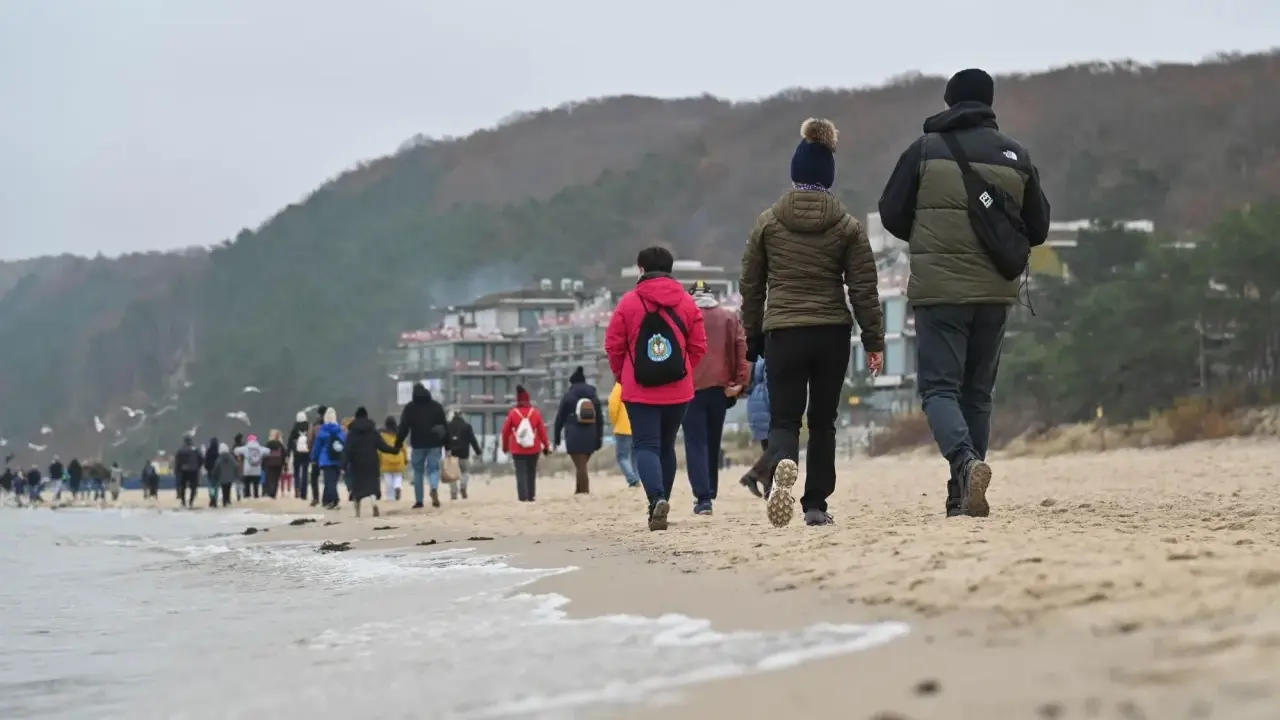 Spacer po plaży w chłodny dzień, idealny na długie weekendy 2026. Grupa ludzi w kurtkach i czapkach idzie wzdłuż morza.