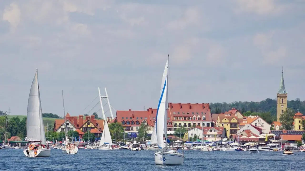 Żaglówki na jeziorze przed malowniczymi mazury miasta z czerwonymi dachami i wieżą kościelną.