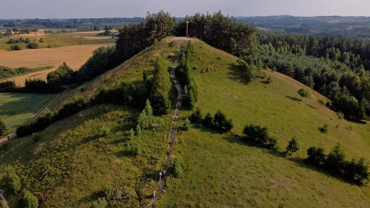 Wzgórze porośnięte trawą i drzewami, ze schodami prowadzącymi na szczyt, gdzie stoi drewniany krzyż. Widok na pola i lasy.