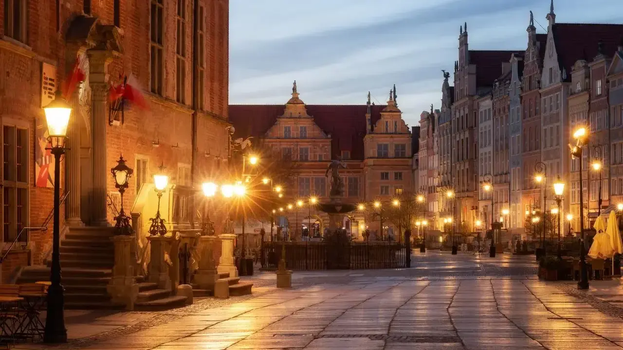 Wieczorny rynek Gdańsk oświetlony latarniami, z fontanną i zabytkowymi kamienicami.