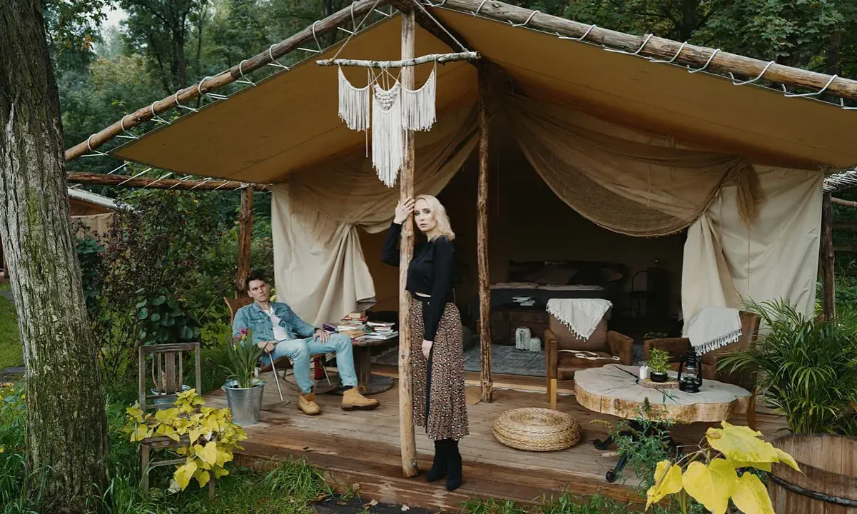 Para młoda relaksuje się w luksusowym kimura glamping namiocie. Mężczyzna siedzi na krześle, kobieta stoi przy drewnianym słupie.
