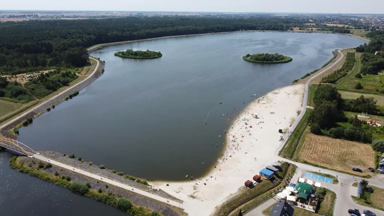 Letnie słońce nad jeziorem opolskim. Ludzie odpoczywają na piaszczystej plaży, pływają w wodzie. W oddali las i zabudowania.