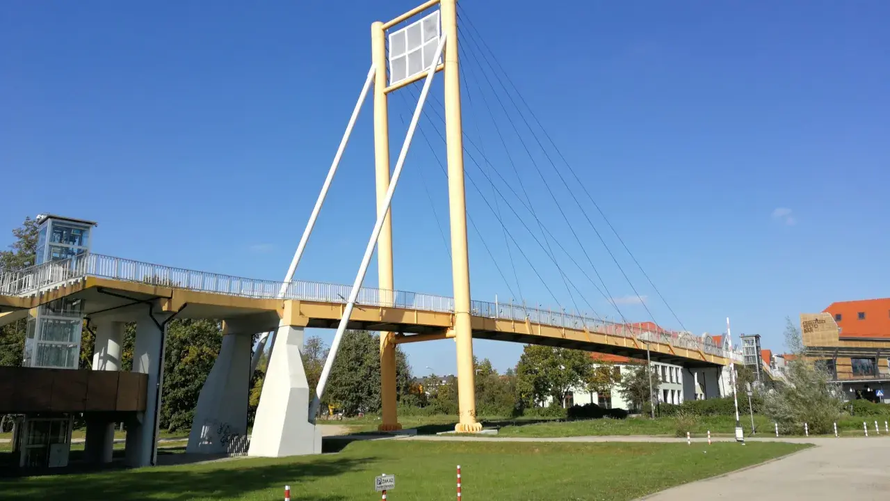 Żółty most pieszy, molo w Giżycku, z charakterystycznymi pylonami i linami, prowadzi do budynków nad jeziorem.