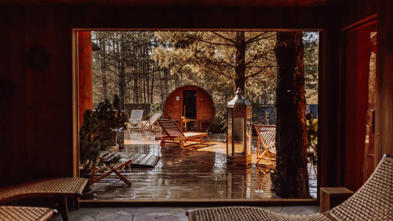 Drewniany domek w lesie, sauna beczka, leżaki i latarnia na mokrym tarasie.