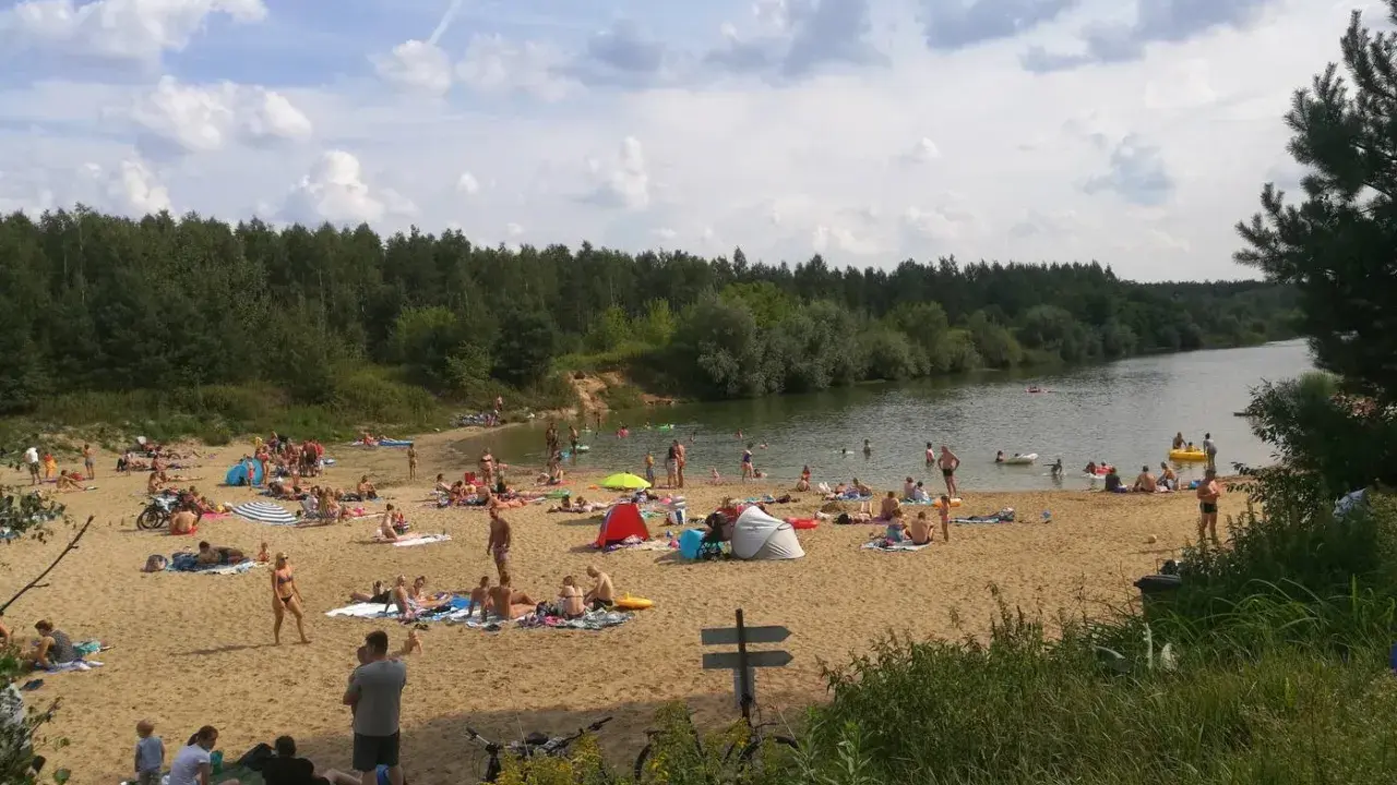 Tłoczna plaża nad jeziorem trzciańskim. Ludzie opalają się, kąpią i odpoczywają na piasku.
