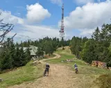 Góra Przehyba z wieżą telekomunikacyjną i schroniskiem. Dwie osoby na rowerach zjeżdżają po kamienistej ścieżce wśród drzew.