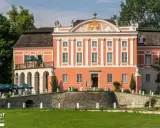 Kurozwęki pałac, z pomarańczową fasadą i białymi zdobieniami, otoczony zielenią. Na pierwszym planie mostek i trawnik.