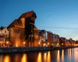 Gdańsk nocą. Historyczny Żuraw i kamienice nad Motławą, rozświetlone latarniami, odbijają się w wodzie.