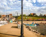Słoneczna plaża nad jeziorem Śniardwy tętni życiem. Ludzie odpoczywają na piasku, kąpią się w wodzie, a obok cumują żaglówki.