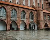 Zamek w Kwidzynie, z ceglaną fasadą i arkadowymi przejściami, w deszczowy dzień. Dwie postacie z parasolem spacerują po mokrym dziedzińcu.