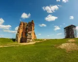 Ruiny ceglanego, opuszczonego zamku na zielonym wzgórzu pod błękitnym niebem z chmurami. Obok wieża w rusztowaniach.