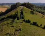 Wzgórze porośnięte trawą i drzewami, ze schodami prowadzącymi na szczyt, gdzie stoi drewniany krzyż. Widok na pola i lasy.
