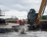 Koparka zrzuca urobek do wody na Zalewie Wiślanym w Elblągu. Widać rozpryski wody i błota.