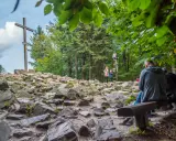 Kamienista ścieżka w **Górach Świętokrzyskich** prowadzi do drewnianego krzyża. Ludzie odpoczywają na ławkach wśród drzew.