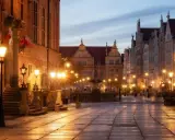 Wieczorny rynek Gdańsk oświetlony latarniami, z fontanną i zabytkowymi kamienicami.