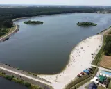 Letnie słońce nad jeziorem opolskim. Ludzie odpoczywają na piaszczystej plaży, pływają w wodzie. W oddali las i zabudowania.
