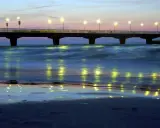 Molo w Kołobrzegu nocą, oświetlone latarniami, odbija się w spokojnych falach morza.