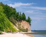 Piękny dzień nad morzem, klif porośnięty drzewami, piaszczysta plaża z spacerującymi ludźmi i statkiem na horyzoncie. Wspaniałe atrakcje nad morzem!