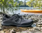 Szare buty sportowe na kamienistej plaży, idealne jako jakie buty na kajaki. W tle pomarańczowy kajak na wodzie.