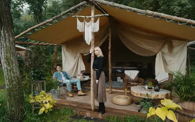 Para młoda relaksuje się w luksusowym kimura glamping namiocie. Mężczyzna siedzi na krześle, kobieta stoi przy drewnianym słupie.
