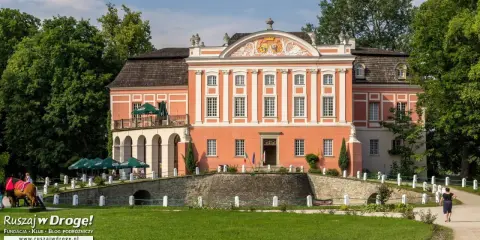 Kurozwęki pałac, z pomarańczową fasadą i białymi zdobieniami, otoczony zielenią. Na pierwszym planie mostek i trawnik.