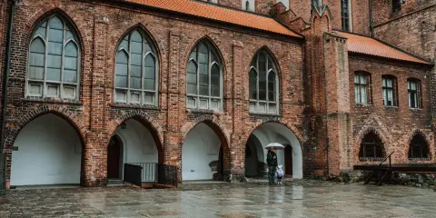 Zamek w Kwidzynie, z ceglaną fasadą i arkadowymi przejściami, w deszczowy dzień. Dwie postacie z parasolem spacerują po mokrym dziedzińcu.