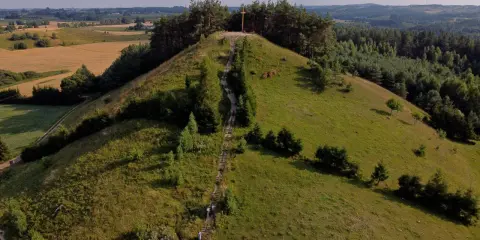 Wzgórze porośnięte trawą i drzewami, ze schodami prowadzącymi na szczyt, gdzie stoi drewniany krzyż. Widok na pola i lasy.
