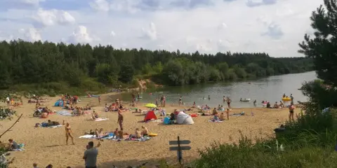 Tłoczna plaża nad jeziorem trzciańskim. Ludzie opalają się, kąpią i odpoczywają na piasku.
