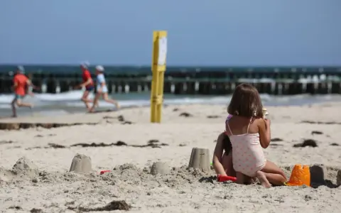 Dzieci budują zamki z piasku na plaży, w tle widać morze i falochrony. Dźwirzyno molo w oddali.