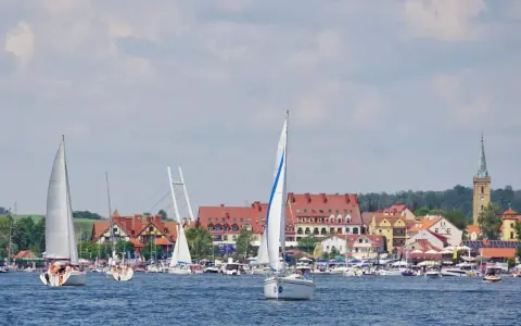 Żaglówki na jeziorze przed malowniczymi mazury miasta z czerwonymi dachami i wieżą kościelną.