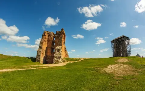 Ruiny ceglanego, opuszczonego zamku na zielonym wzgórzu pod błękitnym niebem z chmurami. Obok wieża w rusztowaniach.