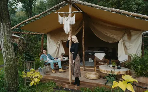 Para młoda relaksuje się w luksusowym kimura glamping namiocie. Mężczyzna siedzi na krześle, kobieta stoi przy drewnianym słupie.