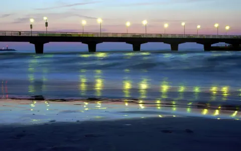 Molo w Kołobrzegu nocą, oświetlone latarniami, odbija się w spokojnych falach morza.