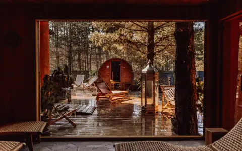 Drewniany domek w lesie, sauna beczka, leżaki i latarnia na mokrym tarasie.
