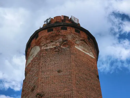 Wieża zamku krzyżackiego w Brodnicy, z cegły, z herbami na szczycie, na tle błękitnego nieba z chmurami.