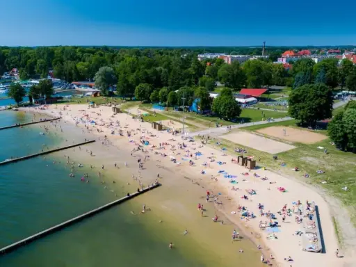 Słoneczna plaża w Giżycku tętni życiem. Ludzie odpoczywają na piasku, kąpią się w jeziorze i spacerują wzdłuż drewnianych pomostów.