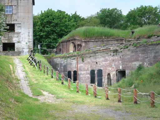 Fort Zachodni w Świnoujściu, zarośnięty trawą i otoczony zielenią, z widocznym starym budynkiem i historyczną artylerią.
