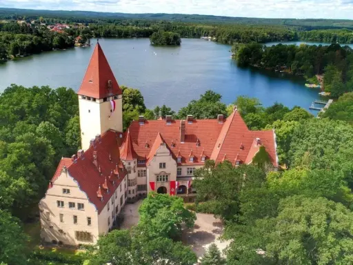 Zamek w Lubniewicach otoczony drzewami, nad jeziorem.