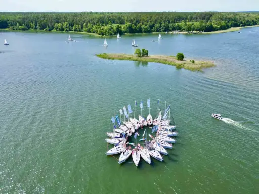 Żaglówki tworzą okrąg na jeziorze, idealne atrakcje Warmia i Mazury. W tle las i mała wyspa.
