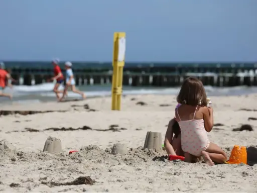Dzieci budują zamki z piasku na plaży, w tle widać morze i falochrony. Dźwirzyno molo w oddali.