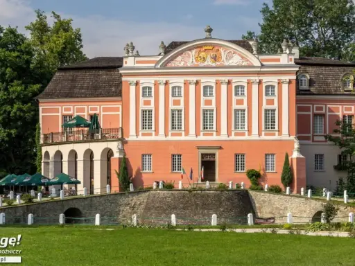 Kurozwęki pałac, z pomarańczową fasadą i białymi zdobieniami, otoczony zielenią. Na pierwszym planie mostek i trawnik.