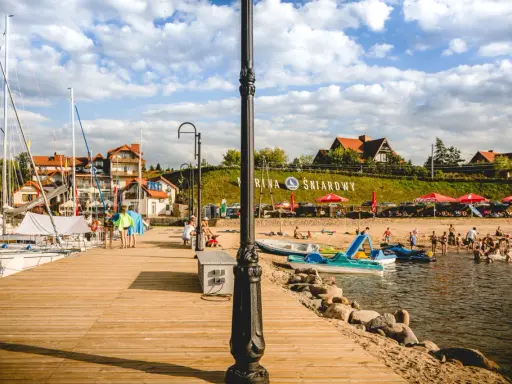 Słoneczna plaża nad jeziorem Śniardwy tętni życiem. Ludzie odpoczywają na piasku, kąpią się w wodzie, a obok cumują żaglówki.